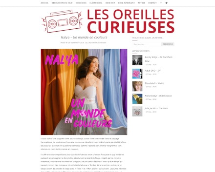Capture d'écran d'un article du site 'Les Oreilles Curieuses' intitulé : 'Nalya - Un monde en couleurs'. L'image principale montre une femme souriante, vêtue d'une robe blanche à bretelles fines, se tenant devant un fond coloré avec des rideaux aux tons orange, bleu, violet et jaune. Le titre de l'article est affiché en rouge, avec le logo du site en haut de la page. À droite, une barre latérale propose des articles similaires.
